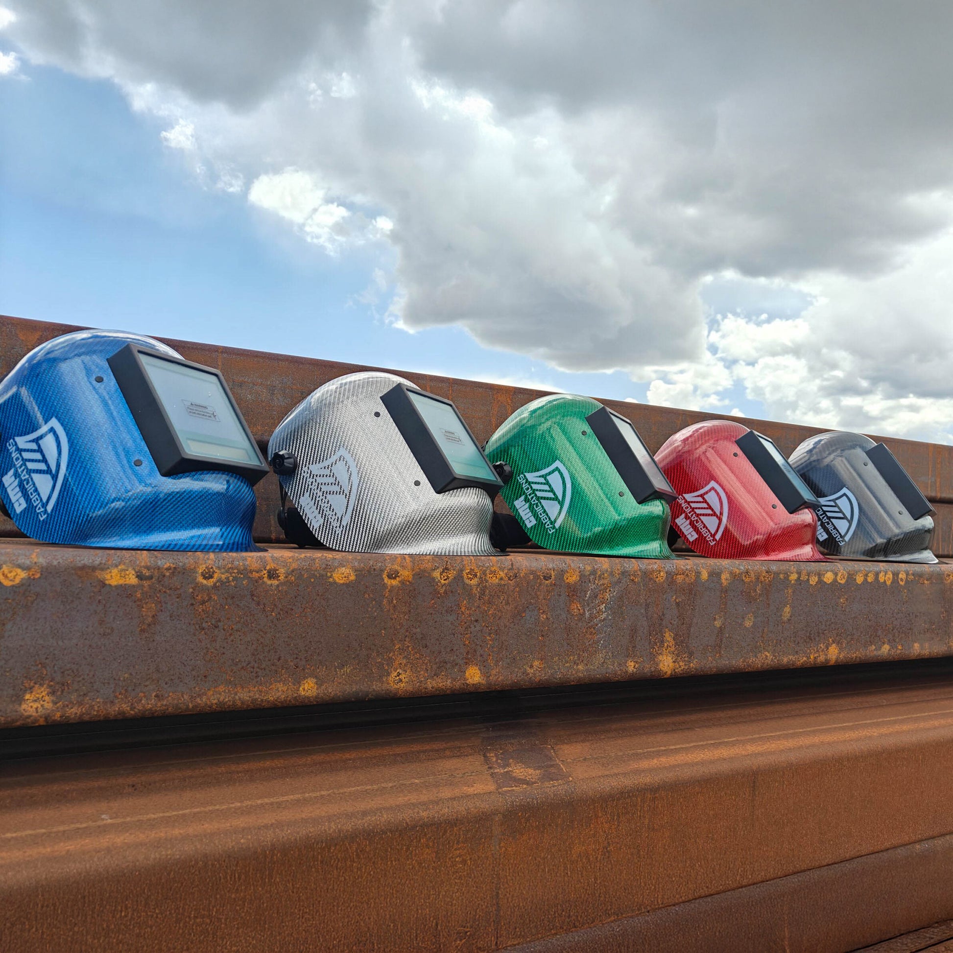 Five colorful CMR Fabrications Pipeliners sugar scoop helmets with logos on a rusty metal surface against a cloudy sky.