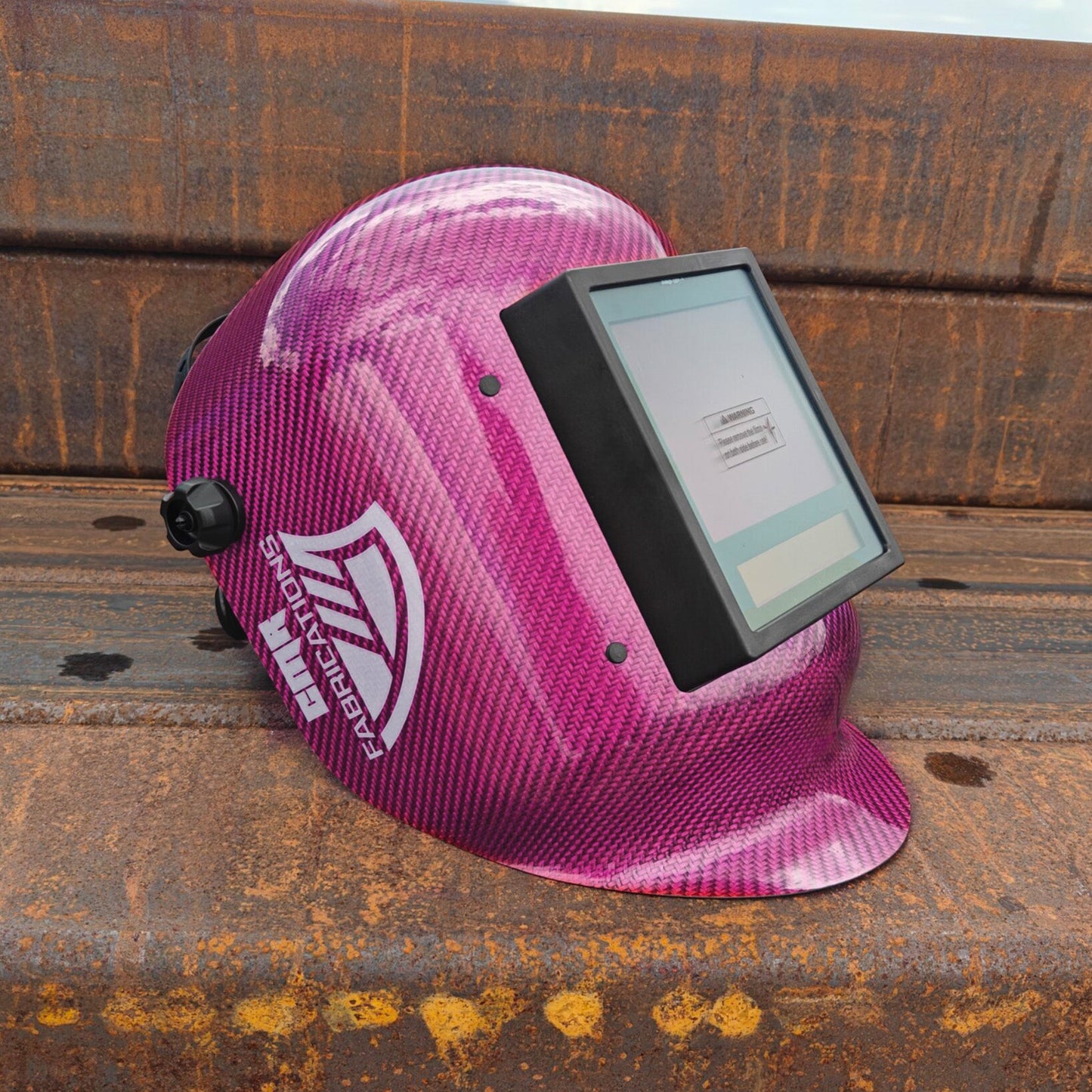 A pink CMR Fabrications carbon-fiber-patterned welding helmet with an integrated dark sun shield, resting on a rusty metal structure outdoors.