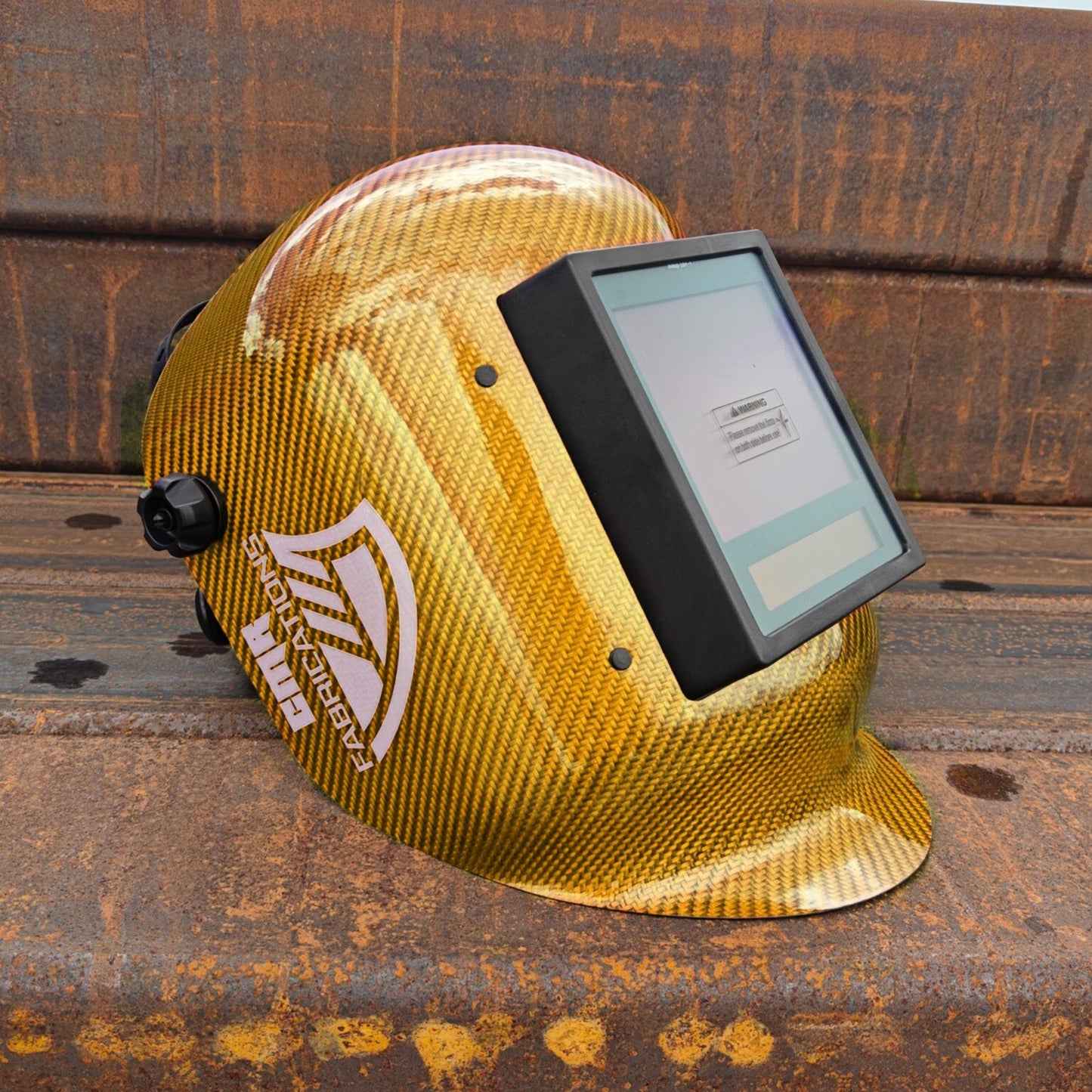 A gold CMR Fabrications carbon fiber patterned welding helmet with a digital screen visor, resting on a rusty metal beam outdoors.