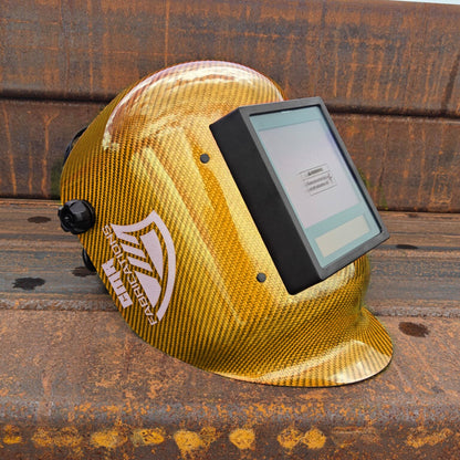 A gold CMR Fabrications carbon fiber patterned welding helmet with a digital screen visor, resting on a rusty metal beam outdoors.